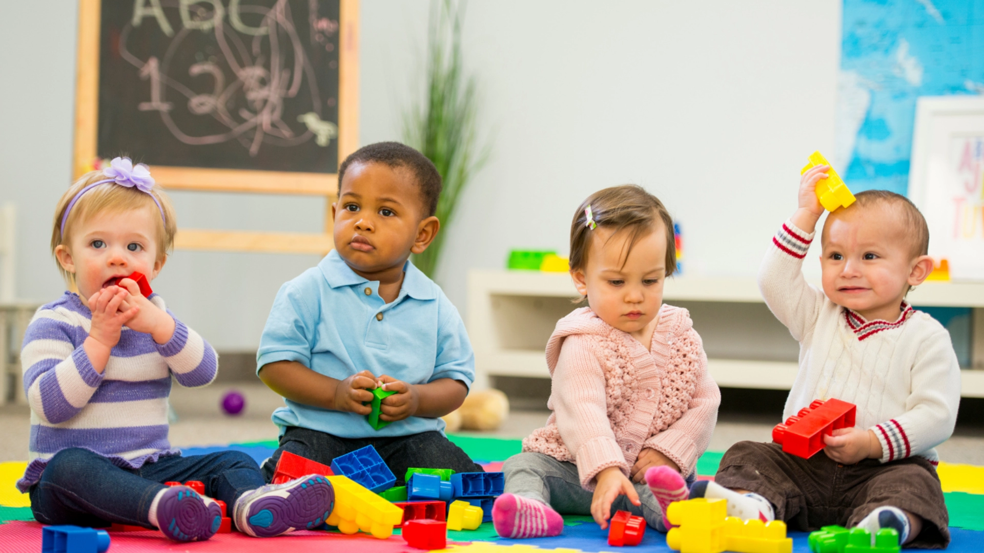 Four toddlers sit on a colorful play mat, engaging with plastic building blocks in a daycare setting. They display various expressions and actions, from chewing a block to raising one playfully. The background includes a chalkboard, a bookshelf, and a world map.