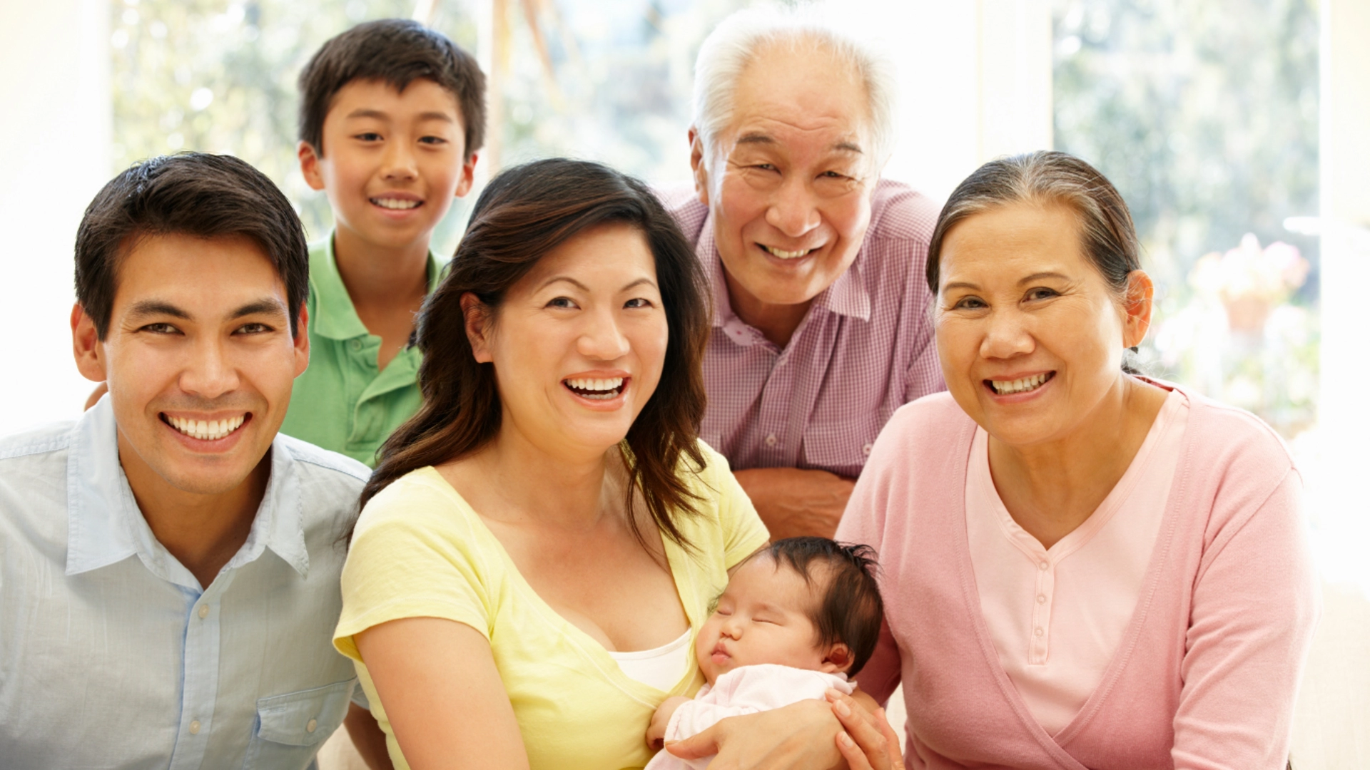 Smiling multigenerational Asian family at home, with a mother holding a sleeping baby, surrounded by loved ones.