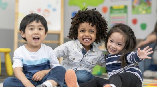 Three young children sitting on a classroom floor, smiling and having fun. One child is wearing a striped shirt, one has curly hair and a white shirt, and one is raising their arm playfully. The background includes educational posters and a whiteboard.