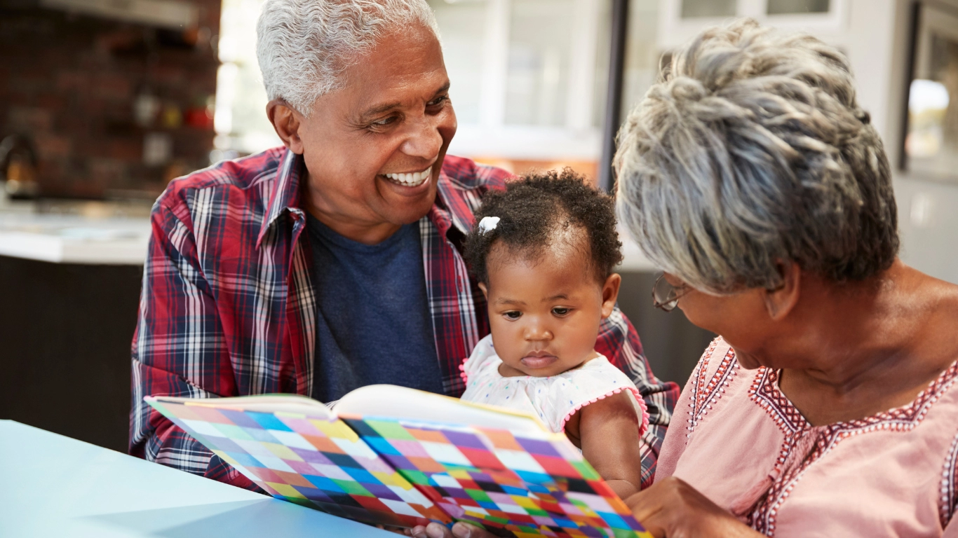 Smiling grandparents reading a colorful picture book with their young granddaughter at a kitchen table, engaging in a bonding moment of care.