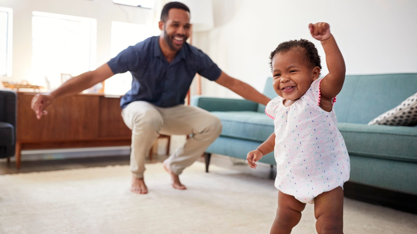 A happy toddler walks toward the camera as a smiling adult encourages them from behind in a cozy living room.