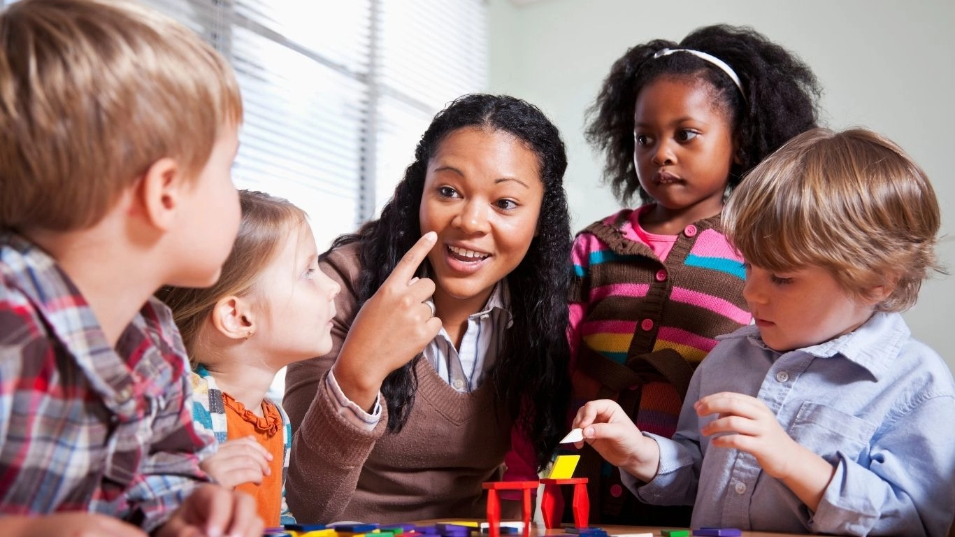 A caregiver engages a group of young children in a learning activity, using gestures and expressions to communicate.
