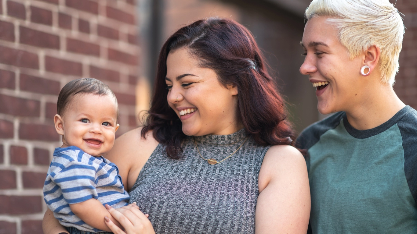 Smiling couple holding their baby outside by a brick wall.