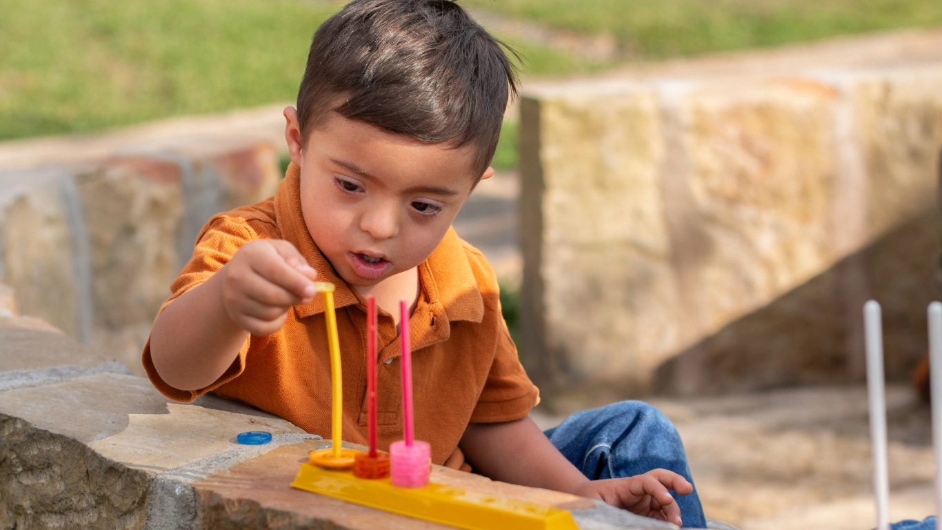A young child with Down syndrome plays intently with a developmental toy outdoors, stacking colorful rings on pegs. The image highlights early learning and developmental support for children with disabilities.