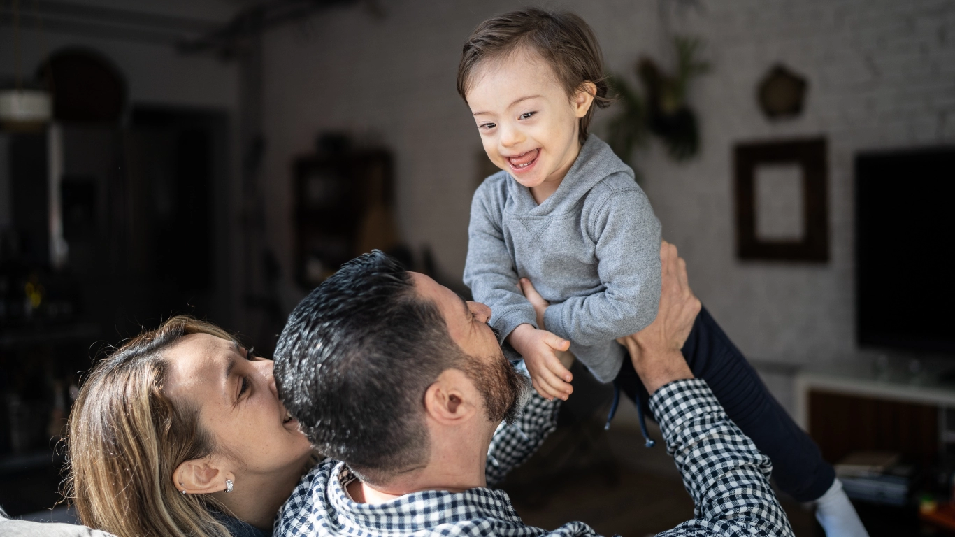 A young child with Down syndrome smiles joyfully as a parent lifts them in the air, while another parent kisses the child on the cheek. The warm, playful moment reflects the nurturing support early intervention programs aim to provide for young children with developmental delays or disabilities.
