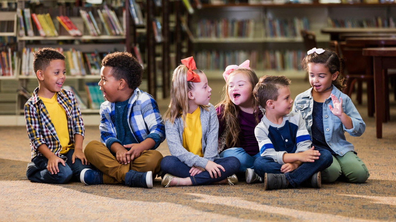 A diverse group of young children sitting in a row on the floor of a library, smiling and interacting with each other. The image represents inclusion, learning, and positive peer engagement in an educational setting.