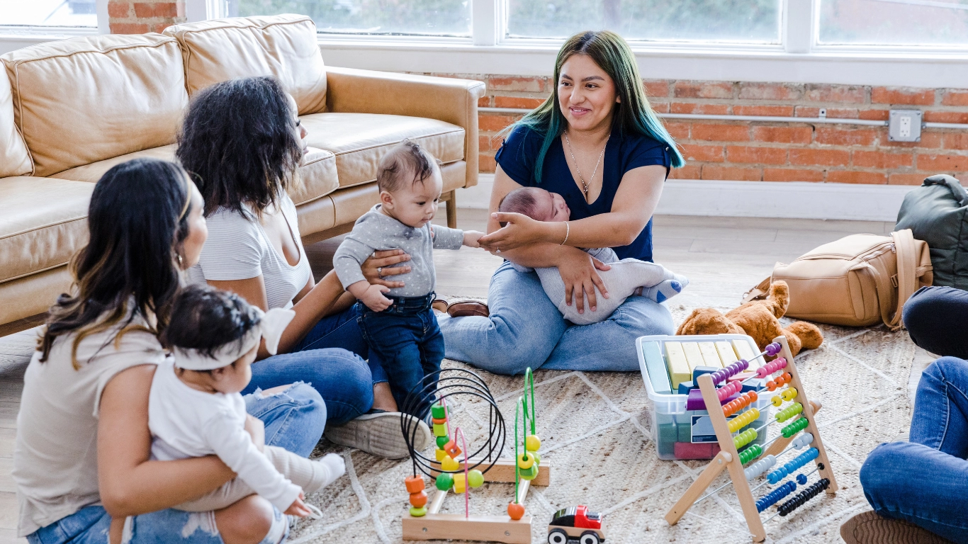 Parents and caregivers sitting together on the floor with young children in a warm, supportive setting. Toys and baby items are visible, reflecting a nurturing, community-centered environment focused on family well-being.