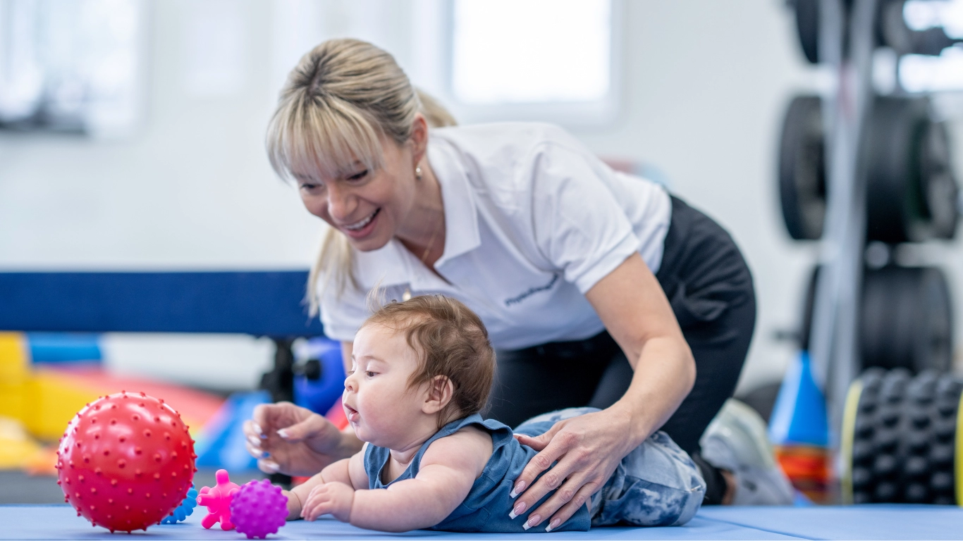 A physical therapist supports a baby with a disability during a developmental play session, guiding them toward colorful textured balls. The scene conveys early support and care for children with developmental needs.