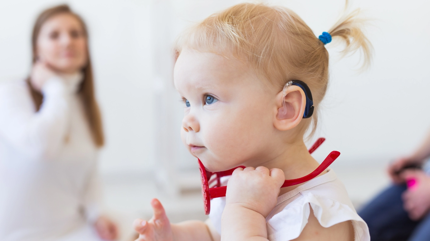 Young child with a hearing aid holds red glasses, highlighting the importance of early identification and support for developmental needs. with a hearing aid holds red glasses, highlighting the importance of early identification and support for developmental needs.