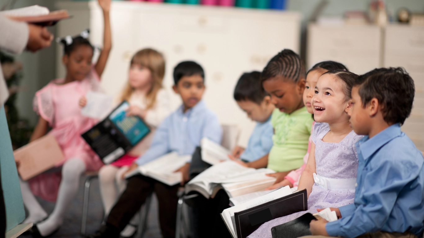 A diverse group of young children sitting in a classroom, holding books and smiling, while a teacher engages with them from the front of the room.