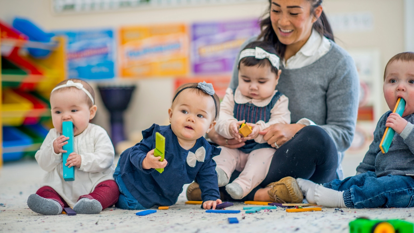 Infants playing with colorful toys on the floor while a child care provider smiles and supervises.