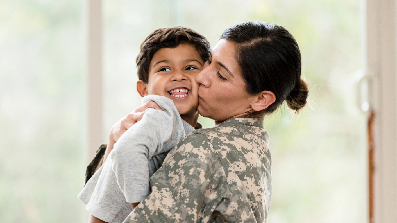 Smiling young boy being held and kissed on the cheek by his mother in military uniform. They are indoors with natural light coming through a window in the background.