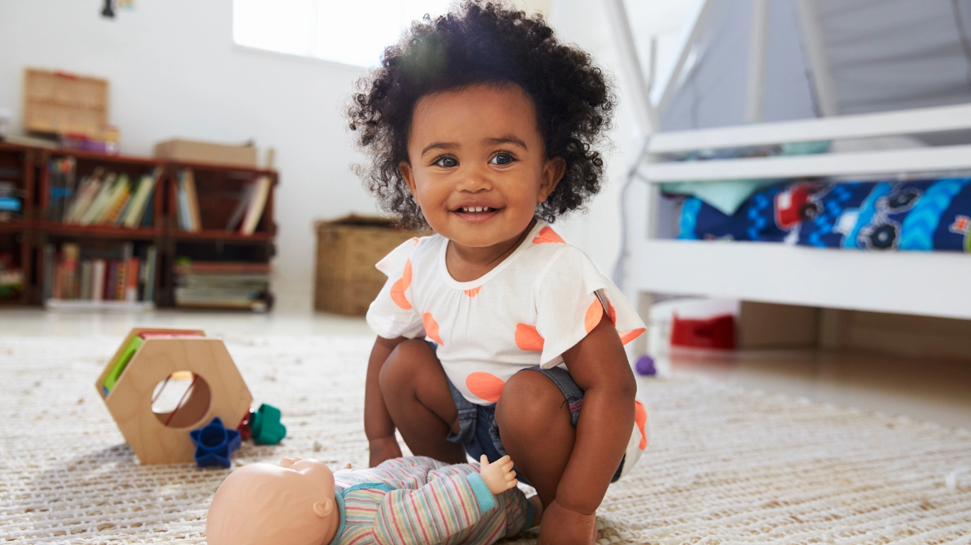 Smiling toddler with curly hair squatting on a carpeted floor in a playroom, surrounded by toys including a doll and a wooden shape sorter. A bunk bed and bookshelf are visible in the background.