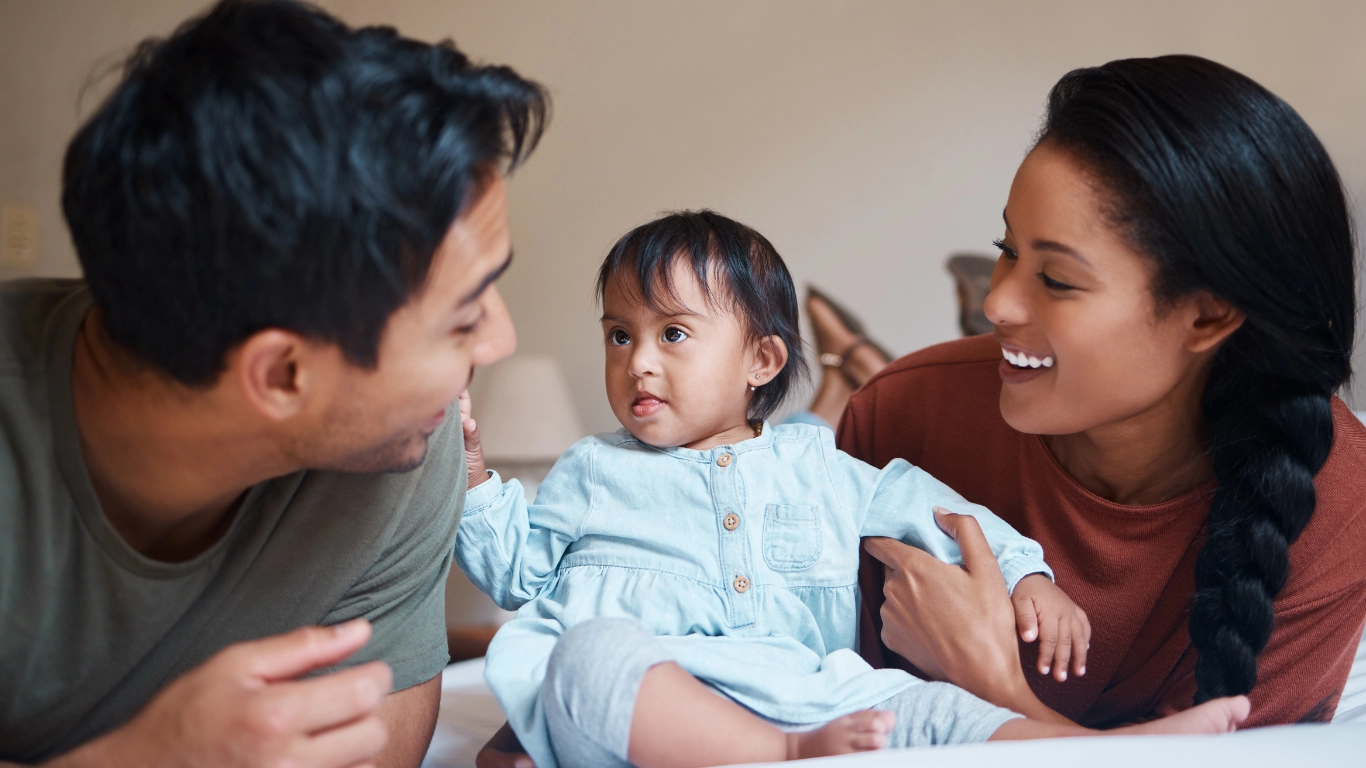 Smiling parents lying on a bed with their baby. The father and mother are looking lovingly at the baby, who is sitting between them and wearing a light blue outfit.