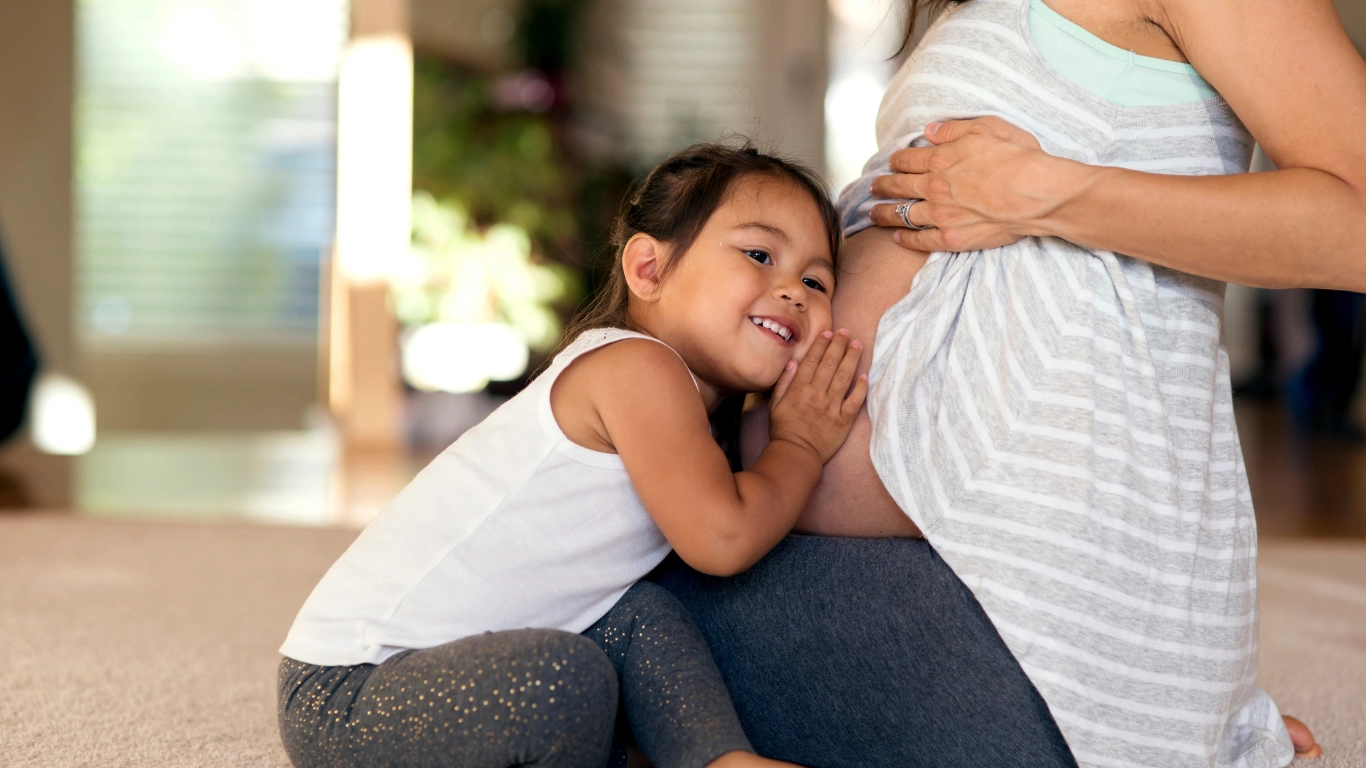A young child smiles with their ear pressed against the bare belly of a pregnant woman, creating a warm and affectionate moment.