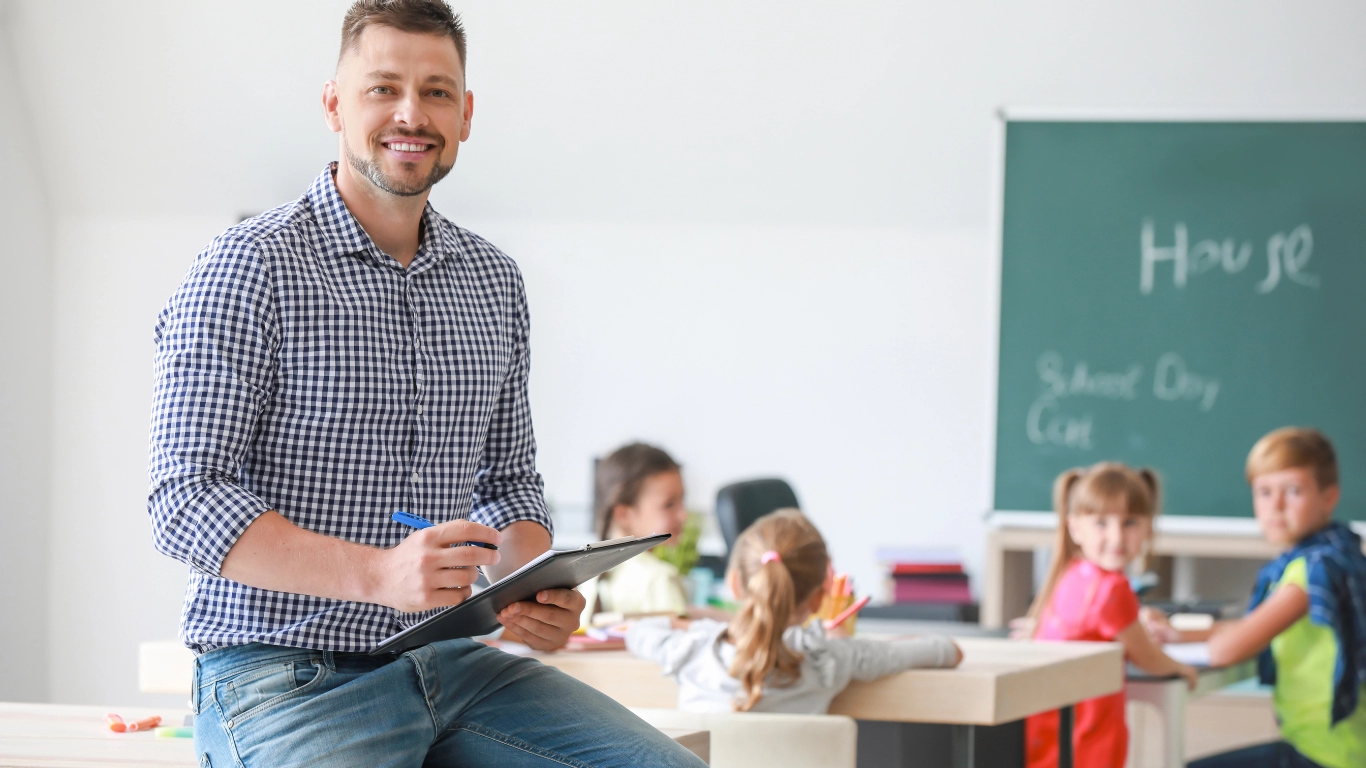 Male educator with clipboard in a classroom with young children and a chalkboard in the background.