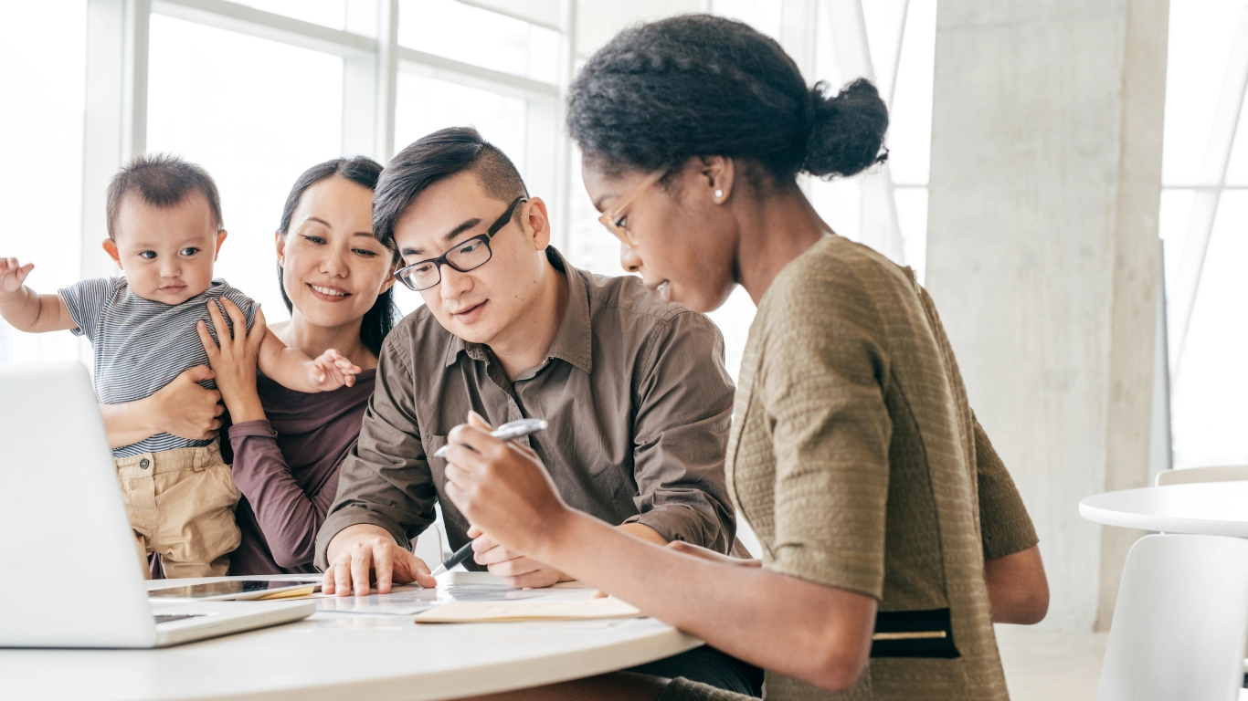 A young family with a baby consults with a financial advisor about child care tax benefits. The advisor explains details using documents and a laptop, while the parents listen attentively, reflecting a supportive and informative discussion in a bright, modern office.