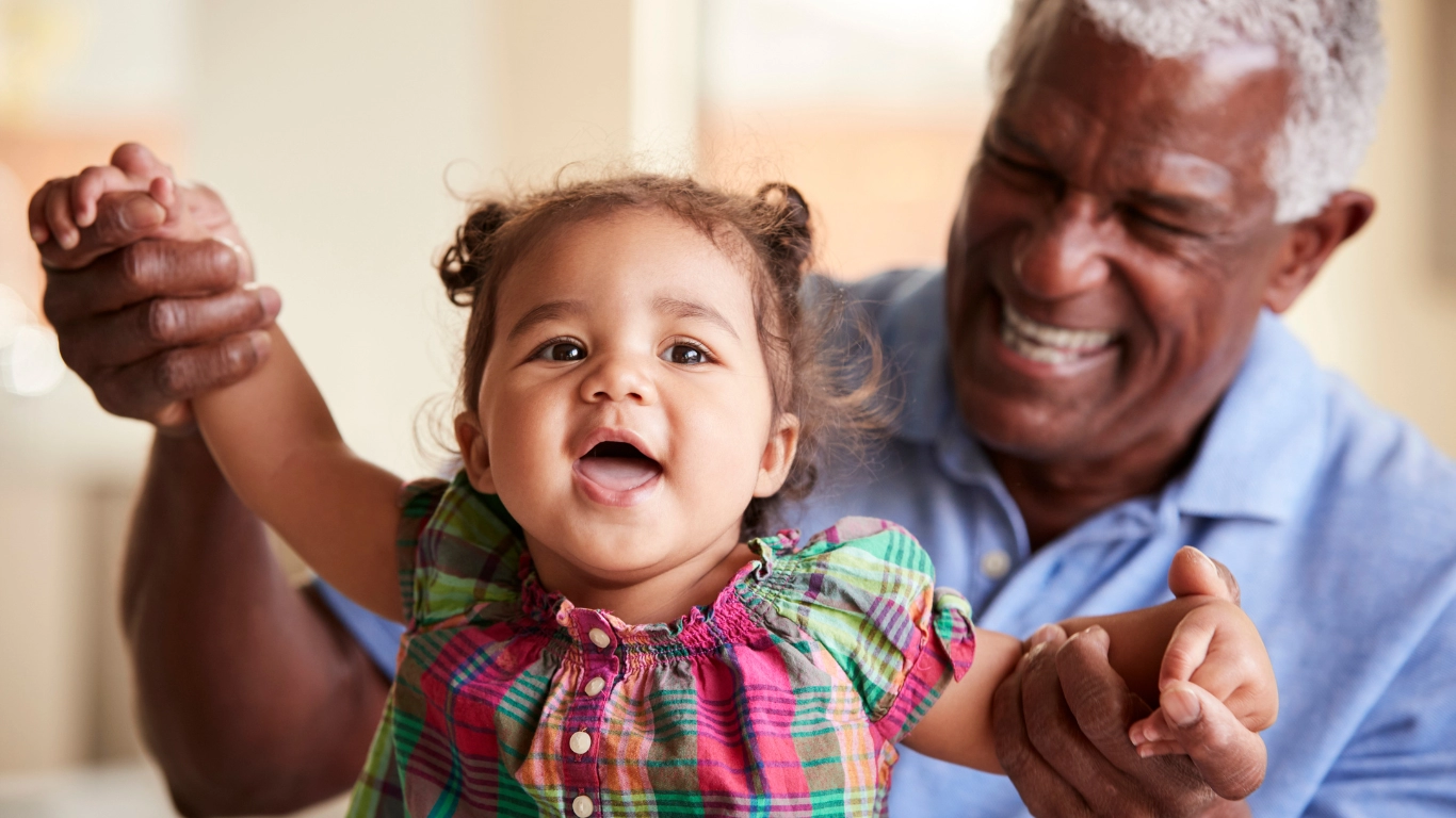 Smiling grandfather joyfully holding up his young granddaughter’s hands as she smiles and looks forward, both sharing a happy moment indoors.